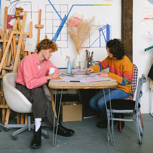 Two artists wearing colourful clothes sit at a table in a studio